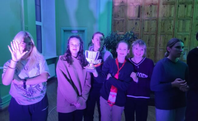 Dažu skolēnu kopbilde, rokās turot kausu, violētā apgaismojumā.