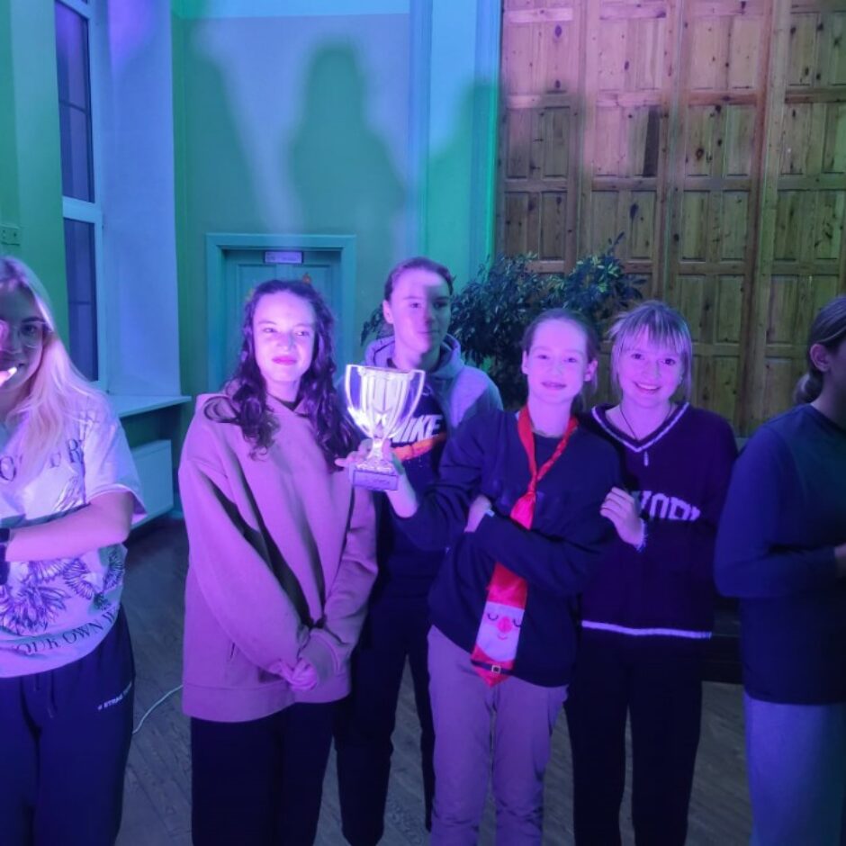 Dažu skolēnu kopbilde, rokās turot kausu, violētā apgaismojumā.