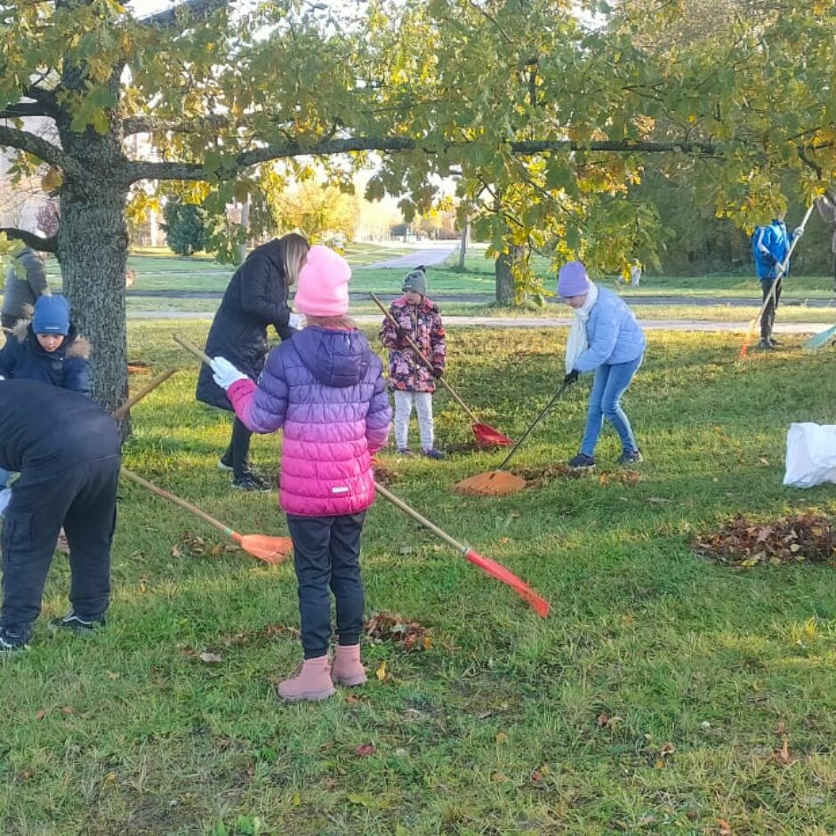 Skolēni uzkopj skolas teritoriju, grābjot lapas.