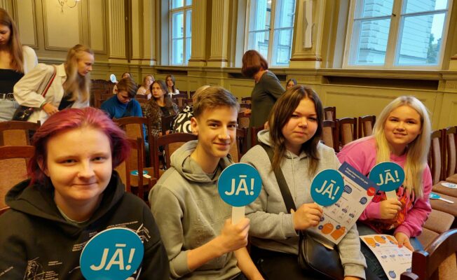 Skolēni apsēdušies Rīgas 2. Valsts ģimnāzijas aktu zālē, rokās turot zilas zīmes ar tekstu 