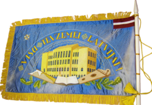 Skolas karoga reverss ar Raiņa ielas skolas ēkas attēlojumu centrā un tekstu "Visu Tēvzemei Latvijai!", zila fona krāsa.