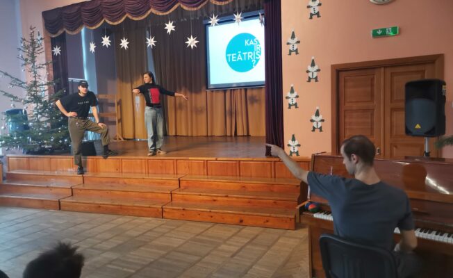 Vīrietis pie klavierēm rāda ar pirkstu uz skatuves esošajiem cilvēkiem