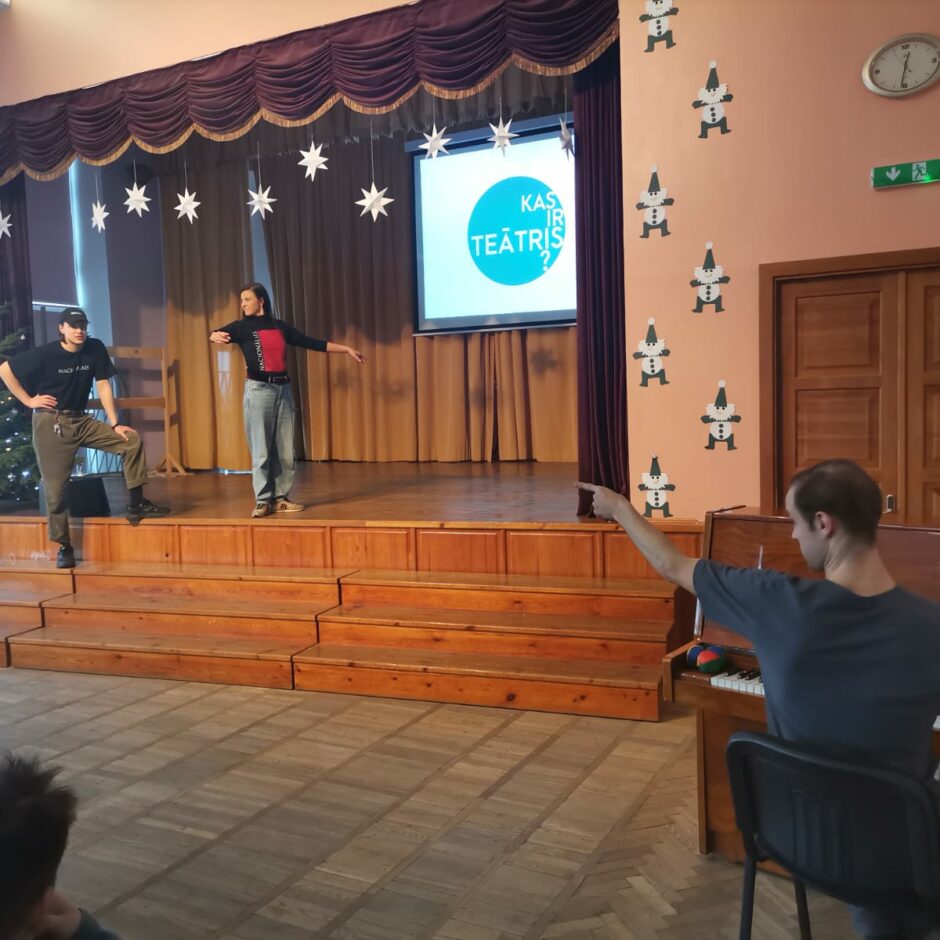 Vīrietis pie klavierēm rāda ar pirkstu uz skatuves esošajiem cilvēkiem